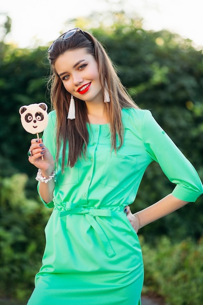 Ritratto di bella ragazza sorridente in abito verde, mostrando alla fotocamera caramelle come panda sul bastone in mano. Donna alla moda con labbra rosse e orecchini lunghi, tenendo la mano in tasca e posando all'aperto.
