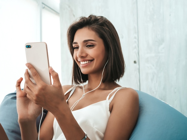 Ritratto di bella ragazza sorridente vestita in pigiama bianco e ascoltando musica con le cuffie. Donna spensierata seduta su una sedia morbida.