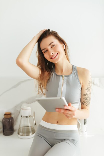 Ritratto di bella signora sorridente in top sportivo in piedi sulla cucina con laptop in mano e auricolari mentre guarda felicemente a porte chiuse