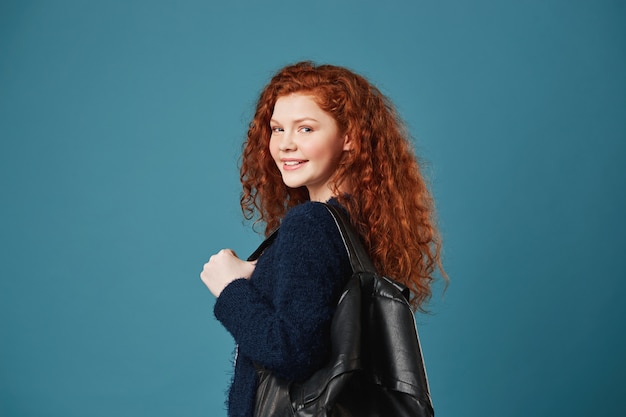 Ritratto di bella studentessa con capelli ondulati rossi e le lentiggini che sorridono con i denti, posando per l'articolo nel giornale della scuola con lo zaino.