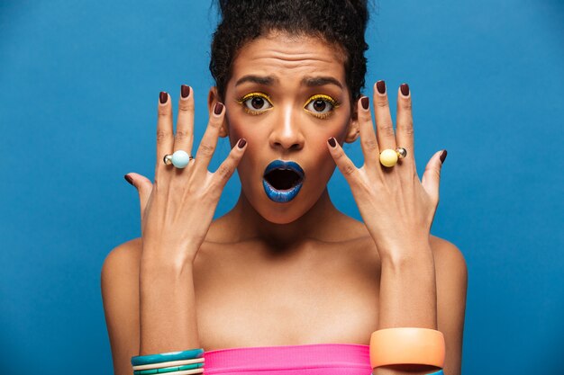 Ritratto di bellezza della donna afroamericana con trucco di modo che dimostra emotivamente gioielli sulle mani che esaminano macchina fotografica, sopra la parete blu