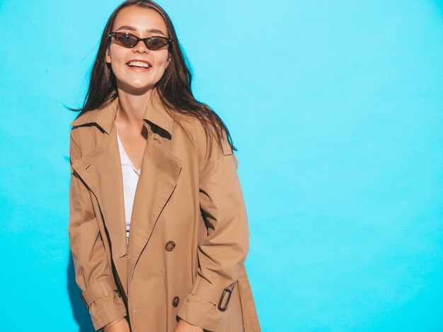 Ritratto di bello modello caucasico della donna del brunette in cappotto ed occhiali da sole marroni. Ragazza che posa vicino alla parete blu