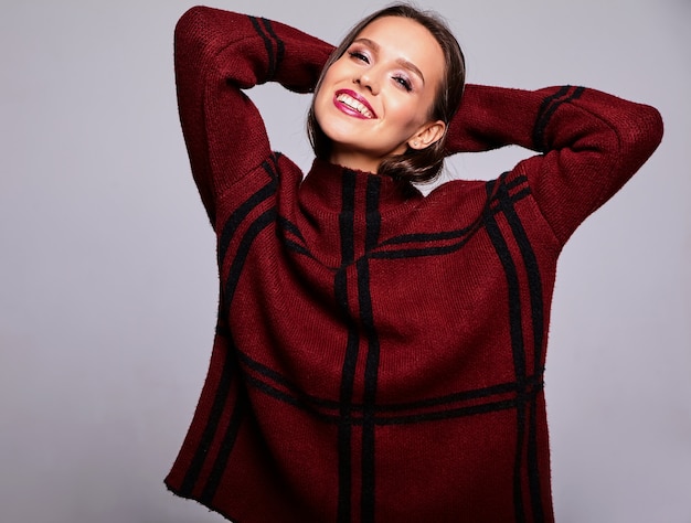 Ritratto di bello modello felice felice della donna del brunette in vestiti casuali del maglione rosso caldo isolati su gray con trucco di sera e le labbra variopinte
