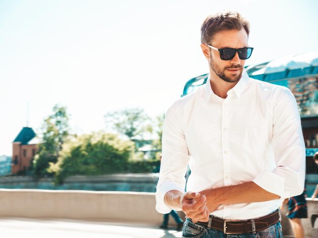 Ritratto di bello sorridente elegante hipster modello lambersexual Uomo moderno vestito con camicia bianca Moda maschile in posa sullo sfondo della strada in occhiali da sole all'aperto al tramonto