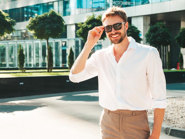 Ritratto di bello sorridente elegante hipster modello lambersexual Uomo moderno vestito con camicia bianca Moda maschile in posa sullo sfondo della strada vicino ai grattacieli in occhiali da sole All'aperto al tramonto