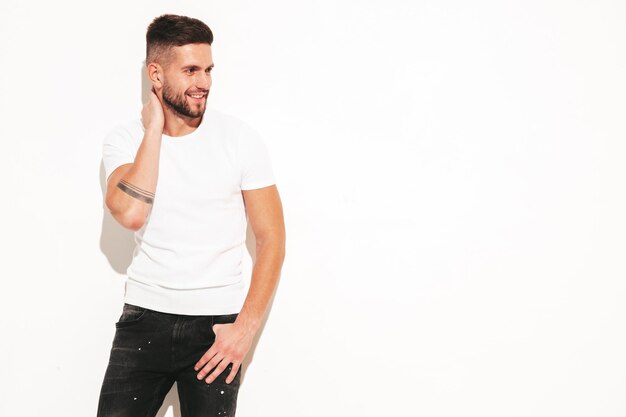 Ritratto di bello sorridente elegante hipster modello lambersexual Uomo vestito con maglietta bianca e jeans Moda maschio in posa vicino al muro in studio isolato