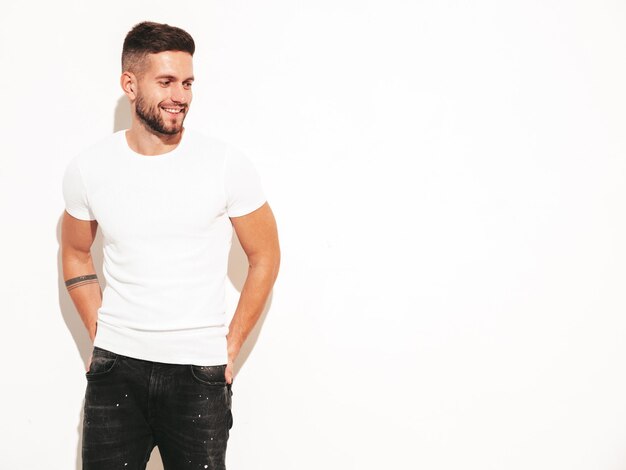 Ritratto di bello sorridente elegante hipster modello lambersexual Uomo vestito con maglietta bianca e jeans Moda maschio in posa vicino al muro in studio isolato