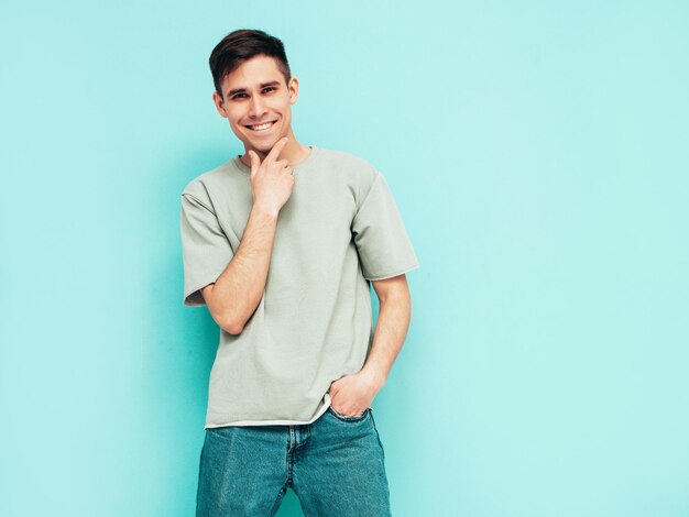 Ritratto di bello sorridente elegante hipster modello lambersexual Uomo vestito con maglietta e jeans Moda maschio isolato in studio in posa vicino al muro blu Allegro e felice