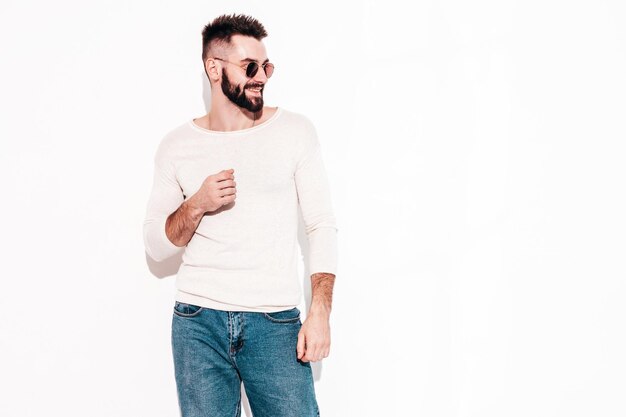 Ritratto di bello sorridente elegante hipster modello lambersexual Uomo vestito con maglione bianco e jeans Moda maschio in posa vicino al muro bianco in studio in occhiali da sole isolati