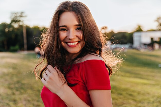 Ritratto di blithesome donna caucasica con i capelli scuri in piedi sulla sfocatura della natura. Ragazza castana felice in vestito rosso che sorride alla macchina fotografica durante il resto nel parco.