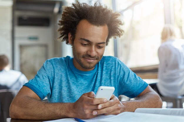 Ritratto di blogger maschio nero con capelli ricci che indossa maglietta casual blu che tiene smart phone moderno utilizzando connessione Internet ad alta velocità, messaggi di testo ai suoi amici seduti al bar