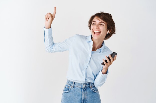 Ritratto di candida donna felice che balla, ascolta musica in cuffie wireless, tiene in mano il telefono e punta verso l'alto, muro bianco