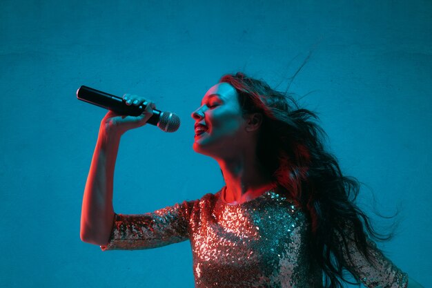 Ritratto di cantante femminile caucasico isolato su sfondo blu studio in luce al neon. Bellissimo modello femminile in abito luminoso con microfono. Concetto di emozioni umane, espressione facciale, annuncio, musica, arte.