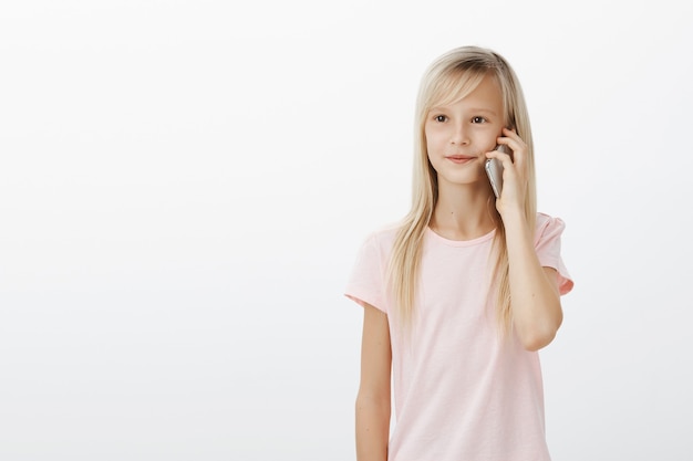 Ritratto di carina giovane figlia con capelli biondi, tenendo lo smartphone vicino all'orecchio e guardando da parte con espressione soddisfatta concentrata, parlando con il fratello