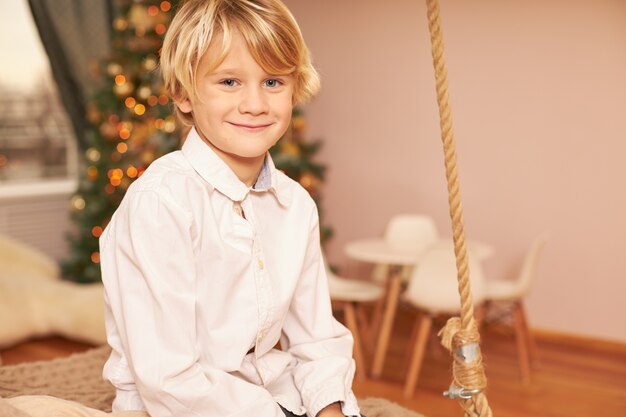 Ritratto di carino ragazzo europeo che indossa una camicia bianca godendo di atmosfera festosa, anticipando la vigilia di Natale, seduto in soggiorno con albero decorato di Capodanno, sorridendo felicemente