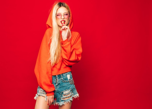 Ritratto di cattiva ragazza giovane e bella hipster in felpa con cappuccio rossa alla moda e orecchini nel naso. Modello positivo che lecca intorno allo zucchero candito