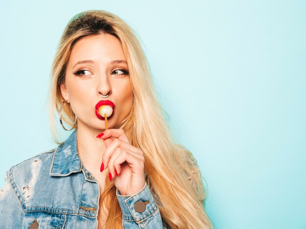 Ritratto di cattiva ragazza giovane e bella hipster in jeans alla moda vestiti e orecchini nel naso. Modello positivo che lecca intorno allo zucchero candito