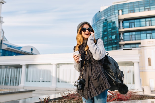 Ritratto di città alla moda di bella ragazza alla moda, camminando con caffè nel centro della città moderna dell'Europa. Gioiosa giovane donna in maglione caldo invernale