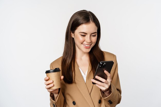 Ritratto di commessa donna aziendale che beve caffè da asporto e legge smartphone guardando mo...