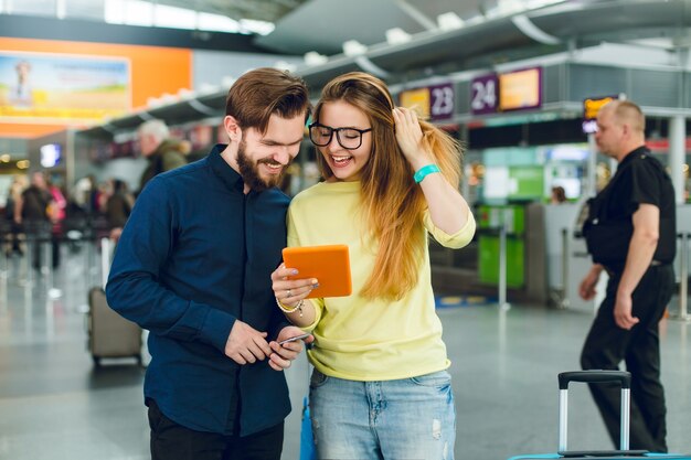 Ritratto di coppia in piedi in aeroporto. Ha i capelli lunghi, occhiali, maglione e jeans. Ha barba, camicia, pantaloni. Stanno cercando su tablet.