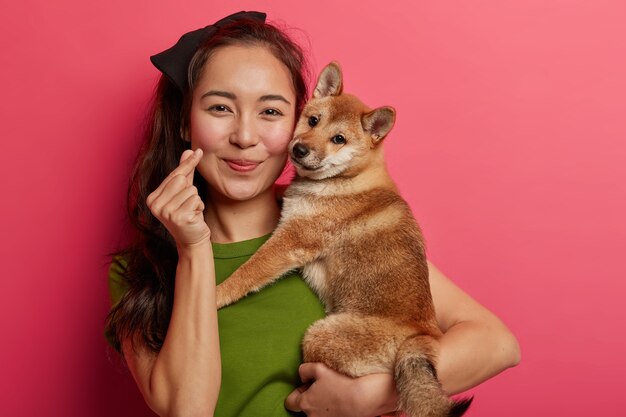 Ritratto di donna abbastanza asiatica mostra il gesto della mano mini cuore, esprime amore, abbraccia cane da caccia di razza giapponese, ama trascorrere il tempo libero con l'animale domestico preferito