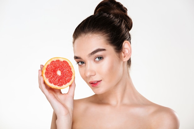 Ritratto di donna abbastanza seminuda con trucco naturale che tiene arancia rossa vicino al suo viso e sguardo