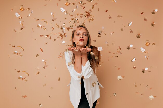 Ritratto di donna adorabile felice invia un bacio sulla parete isolata con coriandoli. Felice celebrazione del nuovo anno, compleanno