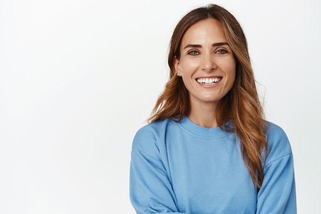 Ritratto di donna adulta ambiziosa e sicura di 30 anni, braccia incrociate e sorridente felice davanti, guardando avanti, in piedi contro il muro bianco