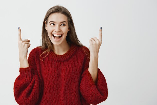 Ritratto di donna affascinante allegra positiva in maglione sciolto rosso, alzando il dito indice e rivolto verso l'alto, guardando da parte con un sorriso felice positivo, in posa per la pubblicità e raccontando di un buon oggetto