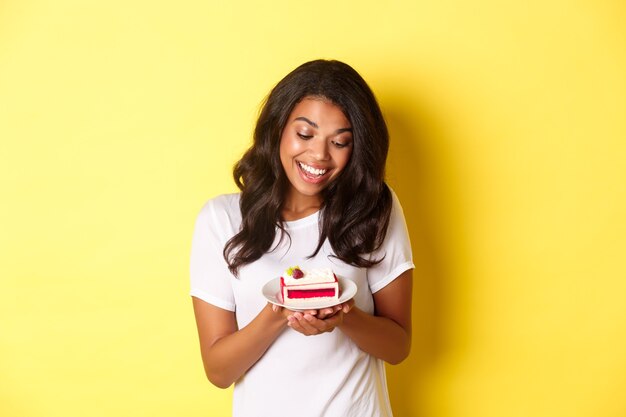 Ritratto di donna afro-americana attraente, guardando un delizioso pezzo di torta e sorridente, in piedi su sfondo giallo