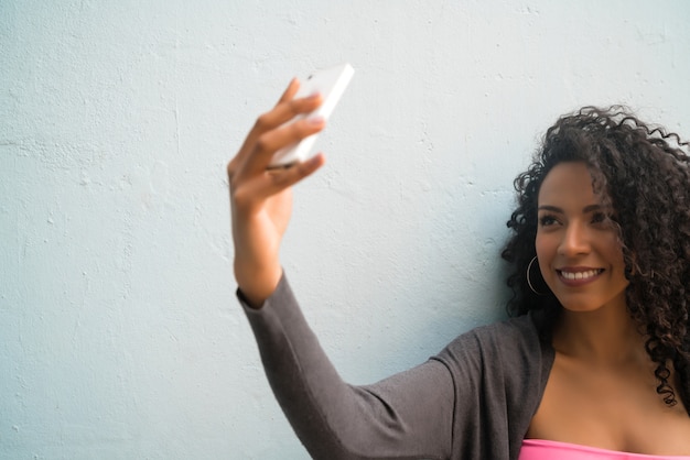 Ritratto di donna afro che cattura selfie con il suo telefono mophile contro il muro grigio. Concetto di tecnologia.