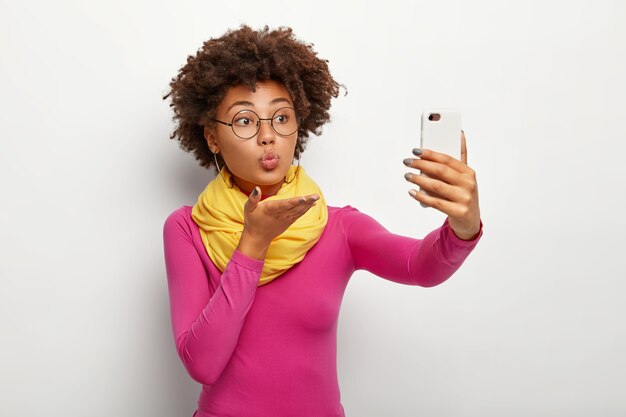 Ritratto di donna afroamericana carina manda un bacio d'aria, prende selfie tramite smartphone, indossa occhiali trasparenti, fa le labbra arrotondate, indossa abiti vivaci, isolato sopra il muro bianco.