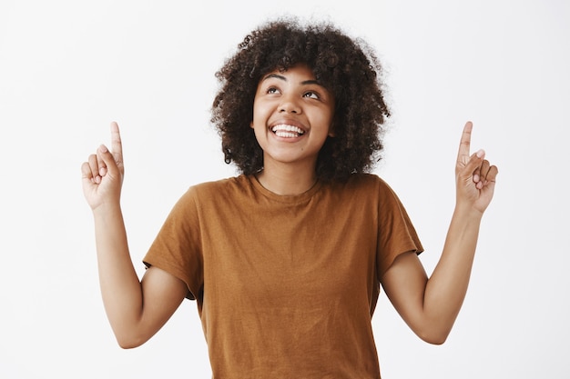 Ritratto di donna afroamericana da sogno attraente in maglietta marrone con acconciatura afro alla ricerca e rivolta verso l'alto con un sorriso gioioso