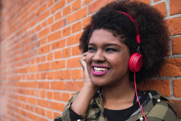 Ritratto di donna afroamericana latina sorridente e ascolto di musica con le cuffie contro il muro di mattoni. All'aperto.