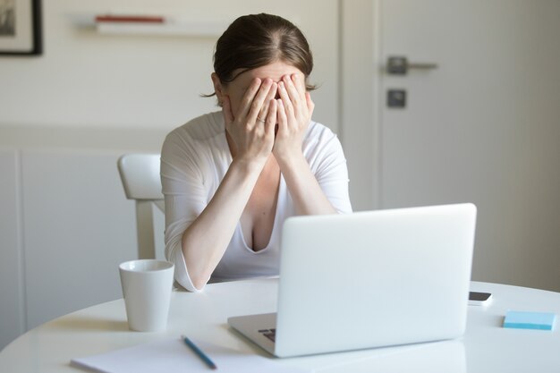 Ritratto di donna alla scrivania con il computer portatile, mani chiudendo la faccia