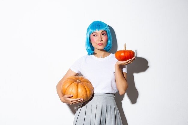 Ritratto di donna asiatica carina premurosa che guarda lontano mentre fa la scelta, tenendo due zucche diverse, decorando la festa di Halloween, indossando la parrucca blu.