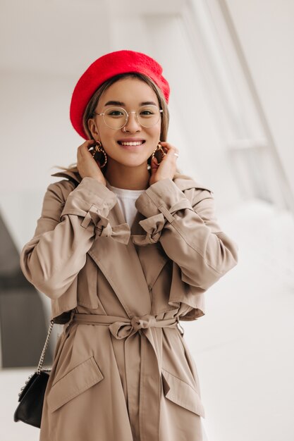 Ritratto di donna asiatica dagli occhi marroni sorridente in occhiali alla moda, berretto rosso in stile francese e trench beige guardando la parte anteriore contro la finestra
