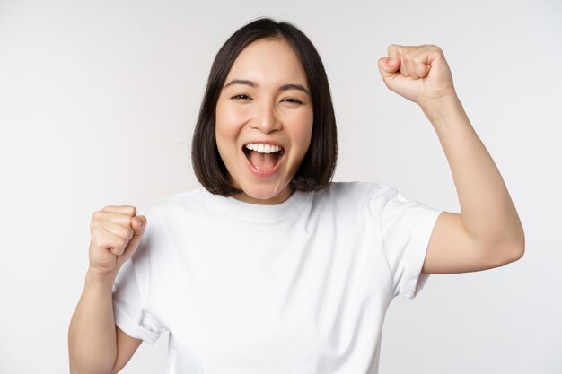 Ritratto di donna asiatica entusiasta che vince celebrando e trionfando alzando le mani per raggiungere l'obiettivo o il successo in piedi su sfondo bianco