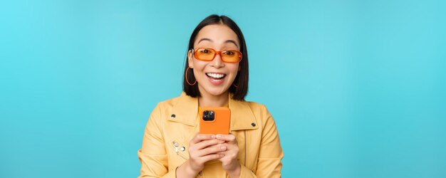 Ritratto di donna asiatica entusiasta in occhiali da sole utilizzando il telefono cellulare sorridente e ridendo guardando felice tenendo smartphone in piedi su sfondo blu