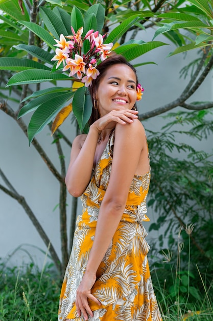 Ritratto di donna asiatica in abito estivo giallo sta con fiore tailandese plumeria nei capelli e orecchini rotondi Donna con trucco leggero fuori sullo sfondo del muro e cespugli verdi