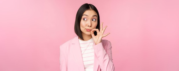 Ritratto di donna aziendale asiatica che mostra il sigillo della bocca chiudere le labbra chiuse sulla promessa del gesto chiave mantenere il segreto in piedi su sfondo rosa in tuta