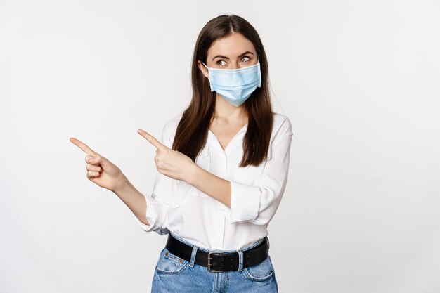 Ritratto di donna aziendale con maschera medica facciale, che punta il dito a sinistra, mostra logo, banner aziendale, in piedi su sfondo bianco.