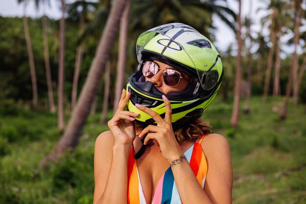 Ritratto di donna bella pilota in casco da motociclista verde giallo e vestito estivo leggero colorato nella giungla sul campo tropicale sotto le palme.