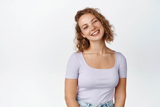 Ritratto di donna bionda attraente con capelli corti ricci, sorridente e guardando felice, in piedi bianco rilassato.