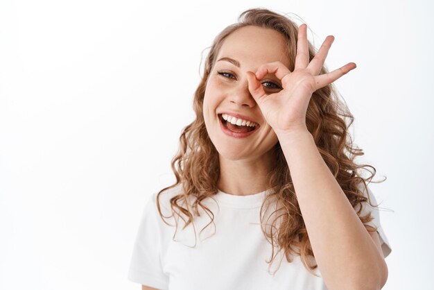 Ritratto di donna bionda con un sorriso schietto naturale che mostra segno giusto dire sì o lodare OK cosa buona in piedi su sfondo bianco