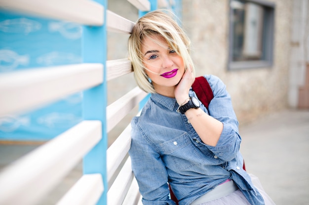 Ritratto di donna bionda sorridente sognante con capelli corti volanti e labbra rosa brillante che indossa camicia di jeans blu