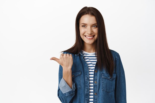 Ritratto di donna bruna allegra sorridente, che punta il dito a sinistra, mostrando il prodotto, lo spazio della copia o il diagramma accanto a lei, in piedi in abiti casual, giacca di jeans, sfondo bianco