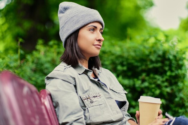 Ritratto di donna bruna con un cappello beve caffè in un parco estivo.