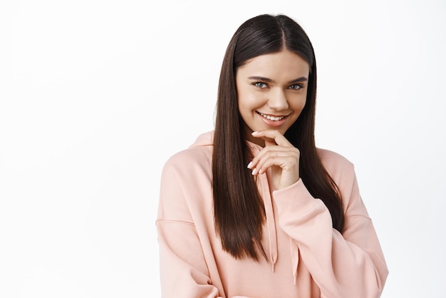 Ritratto di donna bruna pensierosa con lunghi capelli sani e lisci sorridente incuriosito toccando il mento pensieroso e fissando la fotocamera sullo sfondo bianco