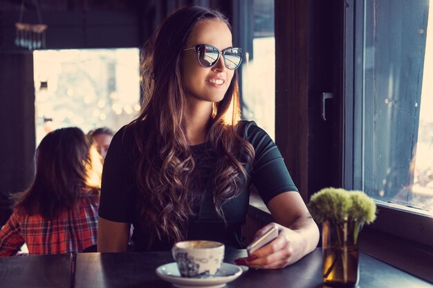 Ritratto di donna bruna sorridente in occhiali da sole, beve il caffè del mattino in un bar.